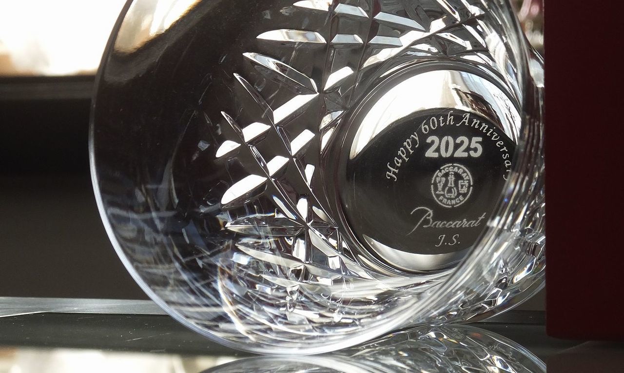 バカラのイヤーグラス、アリア（Aria）タンブラー2025のクリスタルグラスの底面（フット）を捉えた還暦祝い名入れギフトのクローズアップ写真。底面中央には、『Happy 60th Anniversary』という結婚60周年または還暦を祝うメッセージと、その下に『イニシャル』が繊細に名入れ彫刻されているのが鮮明に確認できる。しずくを思わせる繊細なカットを持つグラスの底面に施された、人生の大きな節目を祝う祝福の言葉が、特別でパーソナルな記念品としての価値と輝きを強調している。