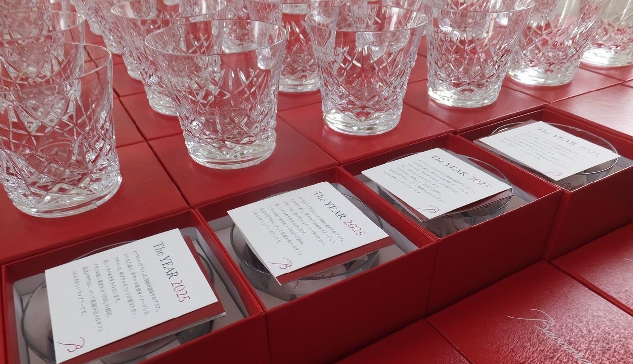 バカラのイヤーグラス、アリア（Aria）タンブラー2025が、多数の真紅のオリジナルボックスの上に整然と並べられている大口注文の集合写真。雪の結晶を思わせる繊細なカットが施されたタンブラーが、鮮やかな赤い箱と透明なクリスタルの輝きの視覚的な対比を生み出している。法人ギフトやイベント記念品として大量に用意されたグラスが、バカラ製品の高級感と供給体制の確かさを象徴する壮観な情景。