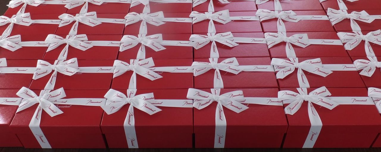 バカラのイヤーグラス、アリア（Aria）タンブラー2025の大口法人注文に対する検品および梱包準備の風景写真。広々とした作業台の上には、多数の真紅のオリジナルボックス（レッドボックス）が山積みされ、その手前で多数のクリスタルグラス（アリア2025タンブラー）が展開されている。スタッフがグラスの傷、名入れ彫刻文字のブレや配置を入念に確認しており、大規模なオーダーにおいてもブランドの品質基準を維持するための徹底した品質管理体制を象徴する情景。