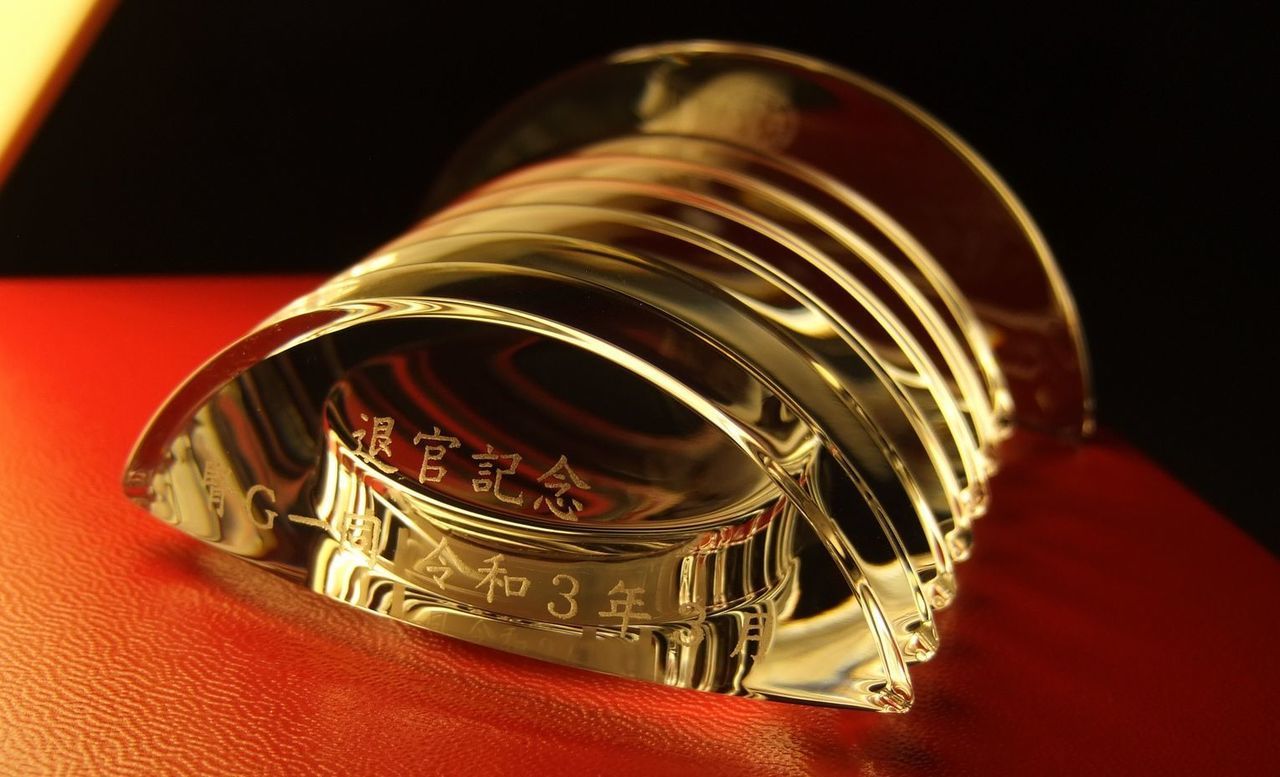 バカラのクリスタル製置き時計『ラランドクロック』の上部に名入れ彫刻を施した退官記念品のサンプル画像。天文学者ラランドの名を冠した円形の星のようなデザインのクリスタル上部に、『退官記念 腎G一同 令和3年』という文字が精巧に刻印されている。透明度の高いクリスタルが周囲の光を反射し、長年の医療・研究への貢献を称えるメッセージを格調高く引き立てており、組織の絆と敬意を伝える特別な贈り物であることを表現している。