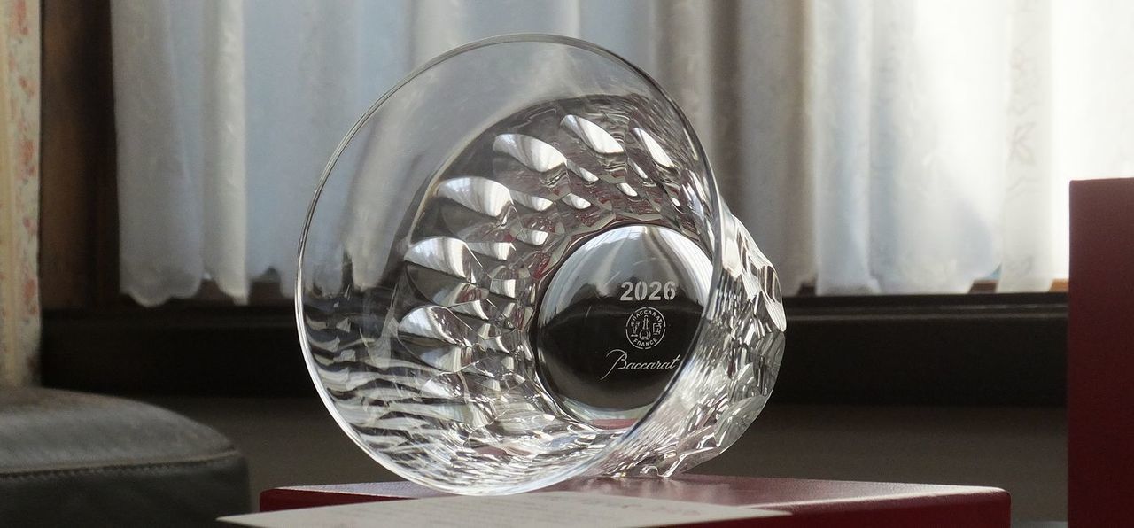 バカラのイヤーグラス、ダリア（Dahlia）タンブラー2026のクリスタルグラスが、真紅のオリジナルボックスの上に横に倒して置かれ、その底面（フット）を捉えたクローズアップ写真。底面の中央には、『2026』という西暦と『Baccarat』のロゴマークが繊細に刻印（エッチング）されているのが鮮明に確認できる。グラスの透明な輝きと箱の鮮やかな赤色が高級感を際立たせ、限定のイヤーグラスであることの確かな証を伝えるプレミアムなギフトの情景。