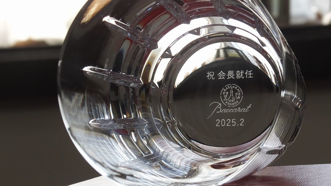 バカラ（Baccarat）のクリスタルタンブラー『アルルカン』の底面に、漢字で『祝 会長就任』の文字と日付が精緻に名入れ彫刻された写真。道化師の衣装に由来する華やかなカットが最高級クリスタルならではの輝きを放ち、長年の功績と新たな重責への就任を祝福する刻印が美しく施され、会長就任祝いや役員就任の御祝にふさわしい最高峰の法人贈答ギフト・記念品のイメージ。