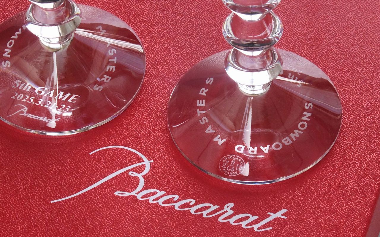バカラ（Baccarat）の『ベガ ワイングラス』の底面に、『スノーボードマスターズ』の大会名、『5th GAME』の回数、および開催期間が精緻に名入れ彫刻されたスポーツ大会記念品の写真。ステム（脚）に連なる幾何学的なダイアモンドカットがモダンな光を放つ最高級クリスタルグラスの底から、大会の歴史と選手の栄光を刻んだ刻印が密やかに美しく輝き、ウィンタースポーツの表彰トロフィーやメモリアルアワードにふさわしい格調高いイメージ。