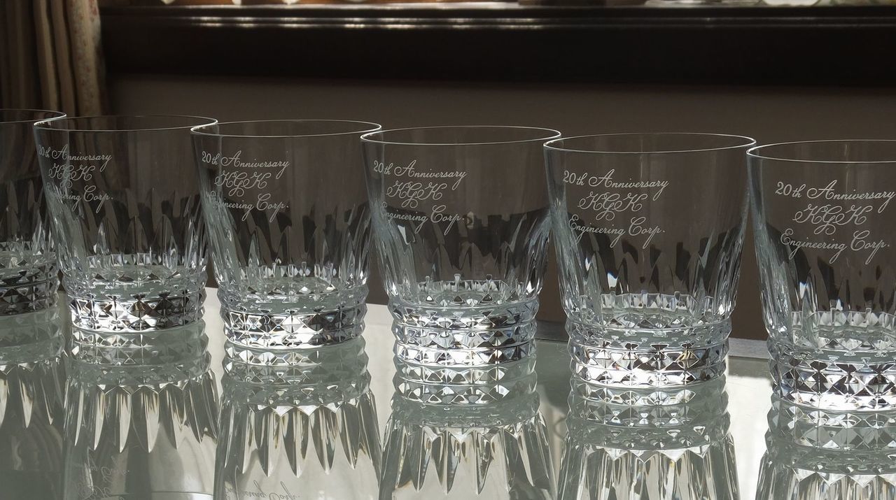バカラの『ピカデリー タンブラー』に創業20周年記念の名入れ彫刻を施した10個のグラスが、モダンなガラステーブルの上に美しく整列している写真。ロンドンのピカデリー・サーカスの名を冠した、伝統的な格子状のカットが特徴的なグラスの側面に、『20th anniversary [企業名]』の文字が、シャープで洗練された英字フォントで精巧に刻印されている。透明なガラステーブルに10客のグラスが放つ幾何学的な光の反射と影が重なり合い、企業の歴史を祝うにふさわしい圧倒的な輝きと重厚な品格を視覚的に表現している。