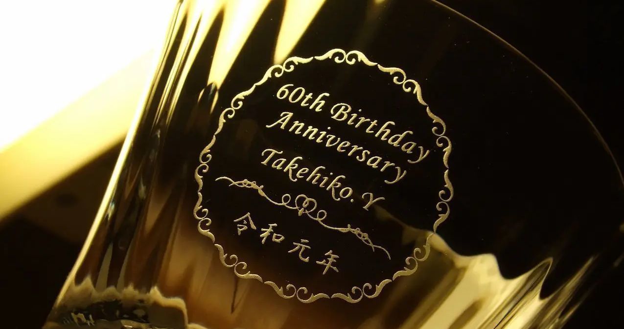 バカラ（Baccarat）の『ミルニュイ タンブラー』の側面に、『60th Birthday Anniversary』のメッセージ、お名前、日付が華やかなオーナメントフレーム（装飾枠デザイン）で優美に囲まれて名入れ彫刻された写真。『千の夜』を意味する放射状の美しく美しいプリーツカットと最高級クリスタルグラスの光に、人生の節目である還暦（60歳）の祝福を刻んだエレガントな刻印が映え、ご家族や大切な方へ贈る最高峰のメモリアルギフトイメージ。