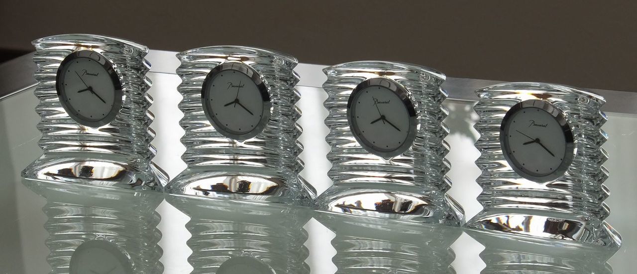 バカラの『ラランド クロック（置き時計）』が、ガラスのテーブルの上に4つ横一列に並べられている様子の写真。ドーム型の重厚なクリスタルと、クラシカルな時計の文字盤を持つクロックが、同じ間隔で整然と配置されている。連続して並ぶクリスタルの輝きは、企業や団体への大口の記念品、または役員への特別ギフトとしての重厚感と高い格式を強調し、時の流れと継続的な成功を象徴的に表現している。