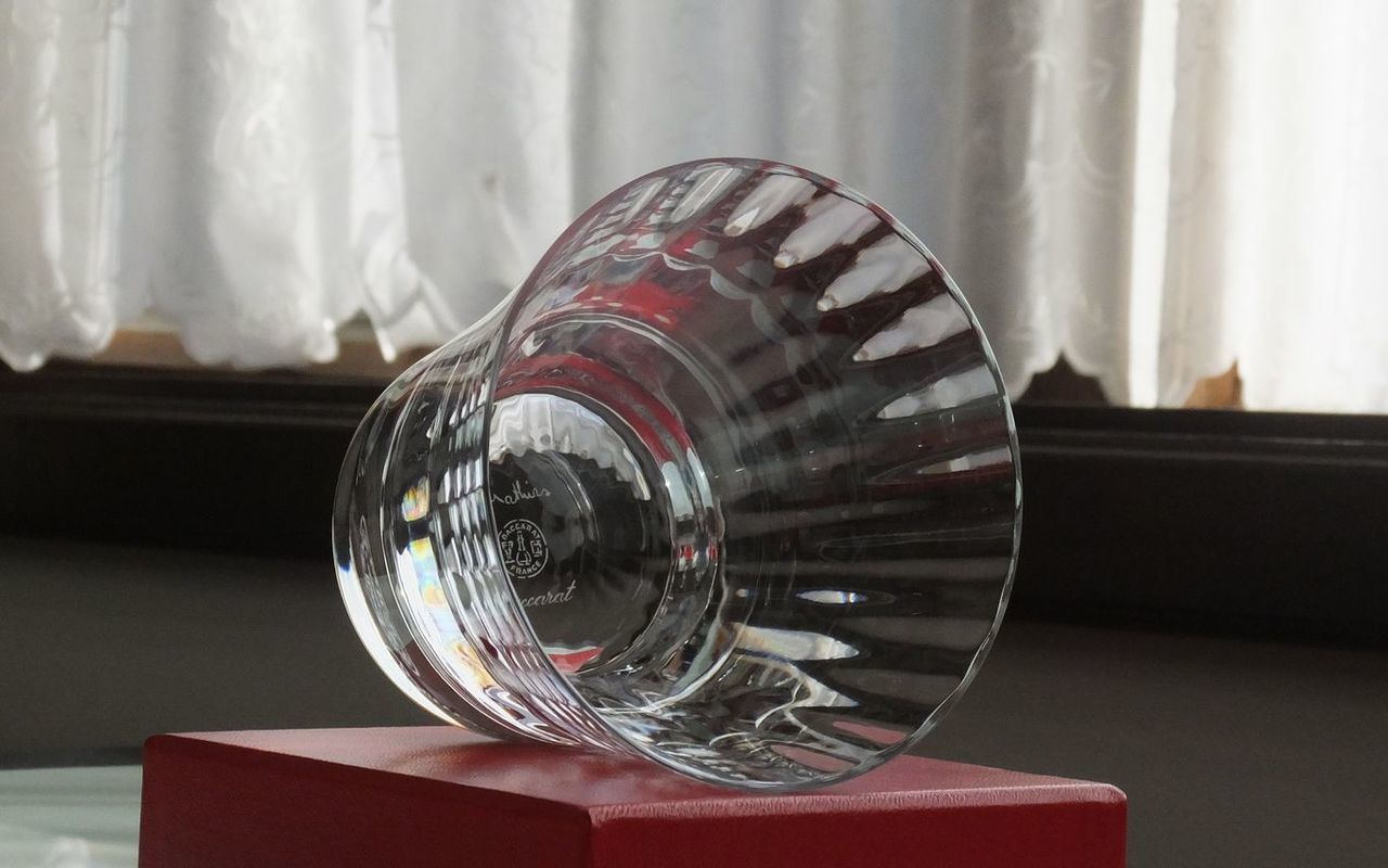 バカラの『ミルニュイ タンブラー』が、バカラのシンボルカラーである赤い純正ボックスの上に、横に倒して置かれている様子の写真。アラビアンナイトをイメージしたミルニュイ特有の優雅なプリーツカットと、深紅のボックスの強烈な色彩のコントラストが際立っている。タンブラーが横向きに置かれることで、そのクリスタルの複雑なカットと輝きがより強調され、最高級ギフトとしての存在感と美しさを視覚的に伝えている。