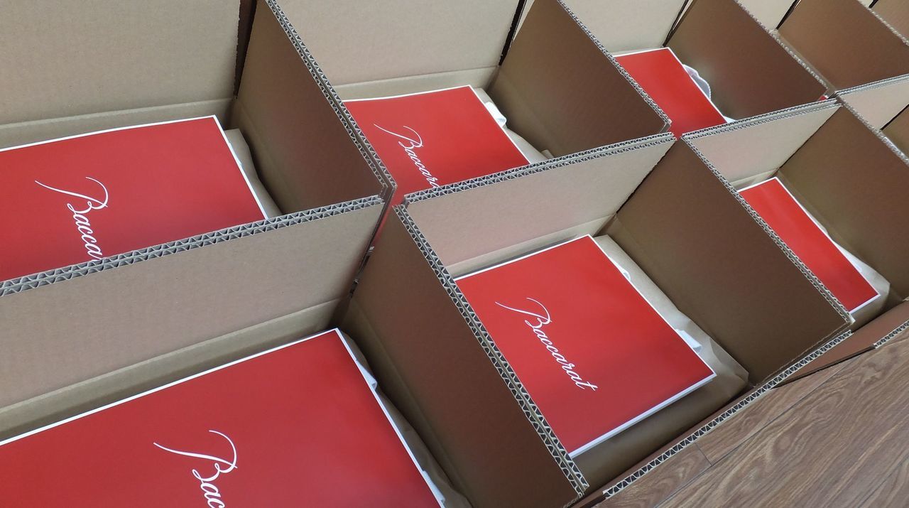 企業の周年記念品として用意されたバカラの2023年イヤーグラス『エクラ』が、大型の段ボール箱の中に80個、均等かつ整然と収められている納品時の様子。一つひとつがバカラの象徴であるレッドボックス**に大切に包まれ、仕切りによって美しく整列している光景は、記念行事に向けた準備の着実さと、80名の受贈者へ向けた感謝の総量を物語っている。2023年モデルならではの『輝き』を意味するカットが施されたグラスが、大切な節目を祝うための万全の態勢で出番を待っている様子を視覚的に表現している。