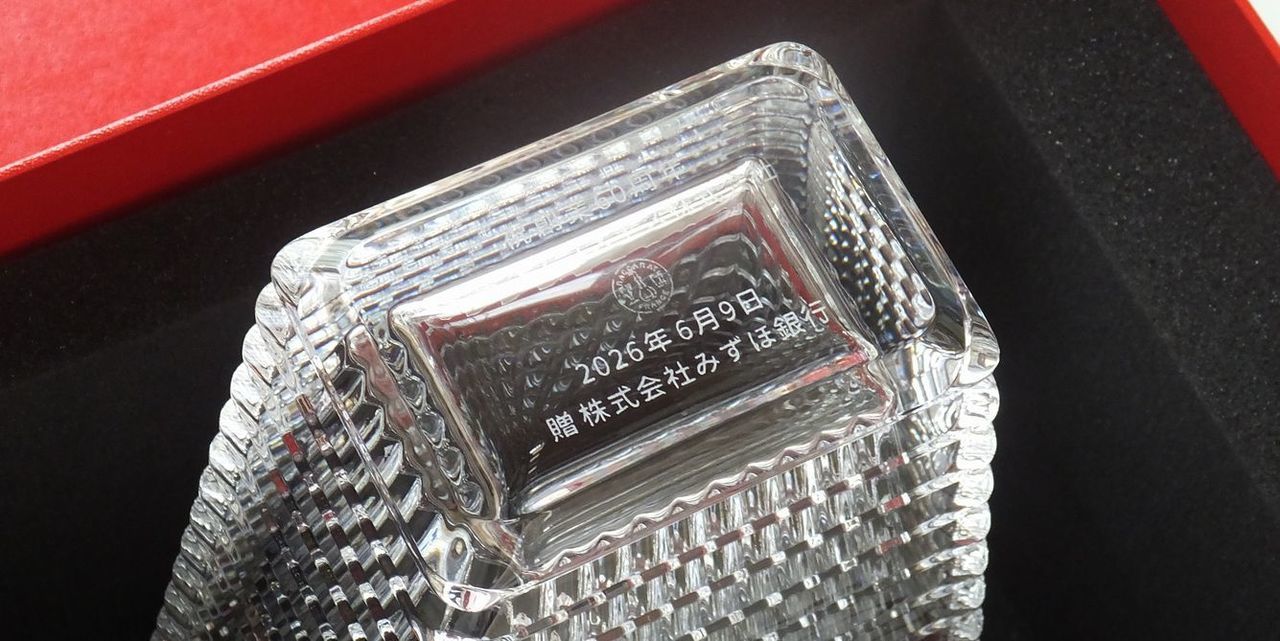 バカラ（Baccarat）の花瓶『アイ ベース スクエア』の底面に、『祝 創業60周年』『企業名』『2026年6月9日』『贈 株式会社みずほ銀行』の文字が精緻に名入れ彫刻された写真。波打つような独特の光の反射と立体的なカットが美しい最高級クリスタルガラス越しに、底面の記念刻印が気品高く浮かび上がり、企業の節目を祝う最高峰の創立周年記念品や金融機関からの贈答品にふさわしい、重厚感と信頼感を備えた格調高いイメージ。