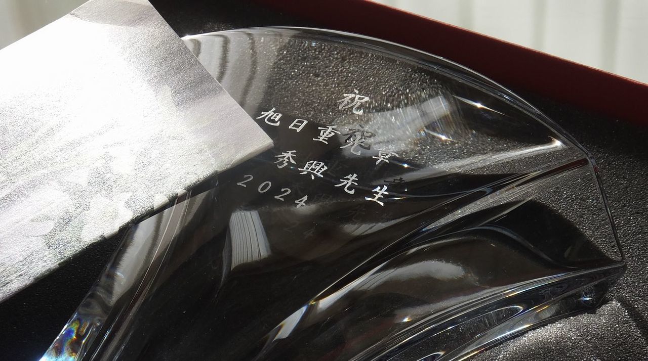 バカラのクリスタル製フラワーベース『ギンコ』の側面に、旭日重光章の受章を祝した名入れ彫刻を施したサンプル画像。扇状の美しいフォルムの中央に、『祝 旭日重光章』『[受章者氏名]』『2024』の文字が、格調高い楷書体で精緻に刻印されている。イチョウの葉をモチーフにしたバカラの名品が、国家的な功労を称える最高の栄誉を象徴しており、長年の献身的な歩みを讃え、その輝きを永遠に留めるための、この上なく優雅で威厳ある記念品であることを視覚的に表現している。