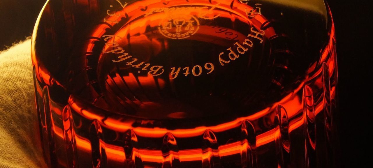 バカラのハーモニータンブラー（Harmonie Tumbler）レッドを暗がりで撮影した還暦祝いの名入れギフト写真。燃えるような深紅のクリスタルが神秘的な輝きを放ち、グラスの側面に刻まれた縦の直線的なカット（ハーモニー）が、わずかな光を捉えて静かに反射している。グラスの底面には、『Happy 60th Birthday [日付] [名前]』という還暦を祝うメッセージが繊細に彫刻されており、長寿と情熱を象徴する特別な記念品の格調高い雰囲気を伝える。