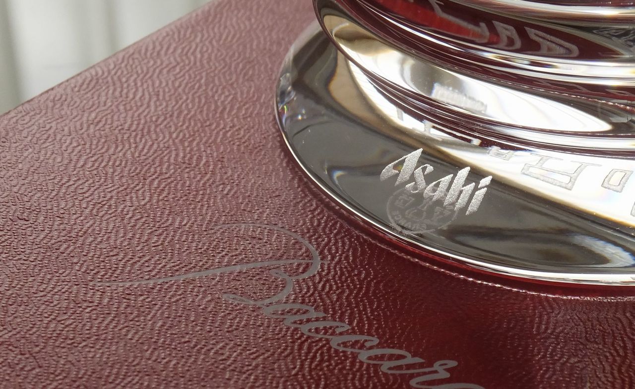 バカラのミルニュイ（Mille Nuits）プレート（皿）のクリスタル部分に、法人ギフトとして『[企業ロゴ] 2024.6.20』という企業ロゴと記念の日付が名入れ彫刻されたクローズアップ写真。ミルニュイ特有のプリーツ状の緻密なカットの中に、企業のブランドアイデンティティと特定の記念日を刻んだメッセージが深く鮮明に刻まれている。世界最高峰のクリスタルの輝きが、事業の成功と大切な節目を末永く記念する格式高い贈答品。