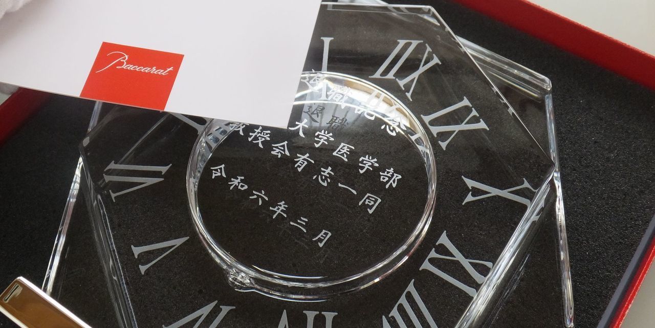 バカラ（Baccarat）の『アビス』クロックの背面に、退職祝いの記念文言、大学名、『教授会有志一同』、および『令和六年三月』の文字が精緻に名入れ彫刻された写真。重厚で建築的なカットが施された最高級クリスタルガラス越しに、背面からの美しい刻印が立体的に浮かび上がり、長年の学術的功績や教育への貢献を称える退職記念品にふさわしい、気品と風格にあふれたイメージ。