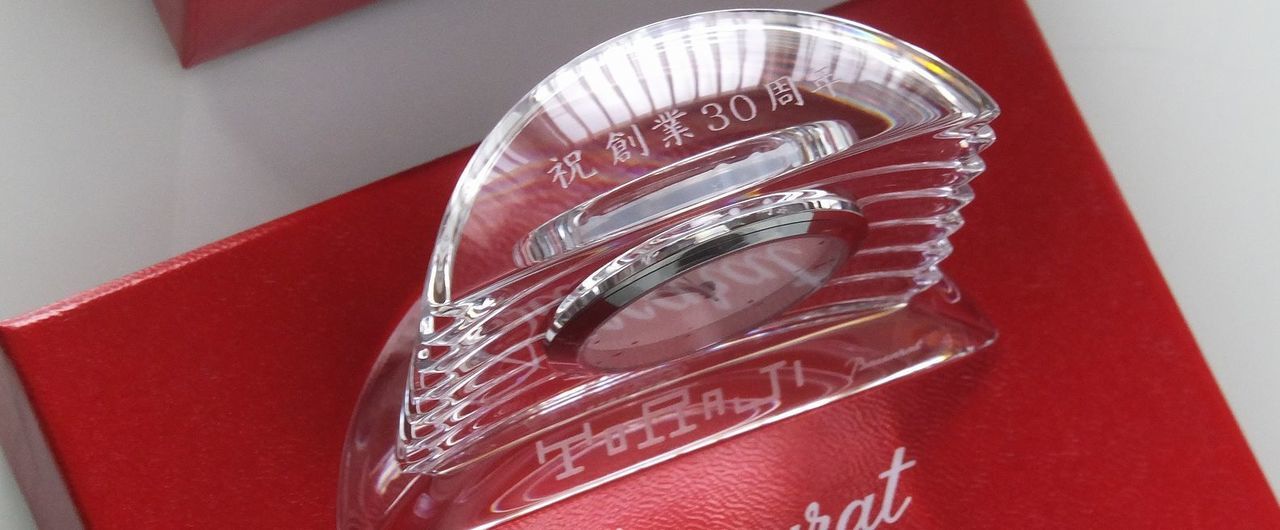バカラの置き時計、ラランドクロック（Lalande Clock）のクリスタル製クロックのクローズアップ写真。アール・デコ調の優美なフォルムと、繊細なダイヤモンドカットが特徴的な時計の上部（カーブしたクリスタル部分）に、『祝 創業30周年 TORAJI』という周年記念のメッセージが名入れ彫刻されている。時間の正確さと不変のクリスタルの輝きが、新たな事業の成功と門出を祝う格式高い記念品