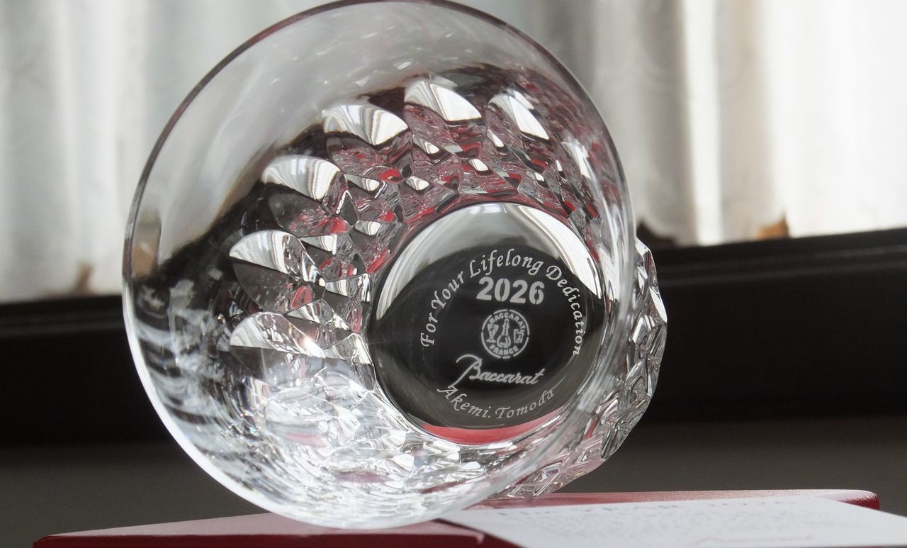 バカラ（Baccarat）の『ダリア タンブラー 2026』の底面に、『For Your Lifelong Dedication』の感謝のメッセージとお名前が精緻に名入れ彫刻された写真。花びら状の美しいカットワークと2026年の年号が刻まれた最高級クリスタルグラスの底から、長年の献身と功績を称える刻印が密やかに美しく輝き、定年退職や永年勤続表彰、特別功労への感謝を伝える最高峰の法人記念品にふさわしい格調高いイメージ。