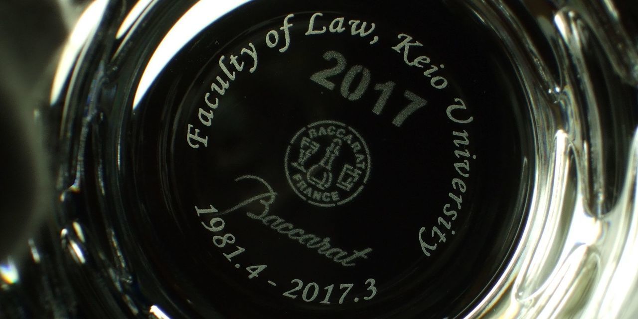 バカラ（Baccarat）の『イヤーグラス 2017』の底面に、『Faculty of Law, Keio University 1981.4-2017.3』の文字が精緻に名入れ彫刻された写真。年号『2017』が刻まれた最高級クリスタルグラスの底面に、慶應義塾大学法学部での長年の在職期間を称える記念英字が美しく浮かび上がり、教授の退職記念や勤続記念、学術的功績を称える特別で格調高いギフトにふさわしいイメージ。