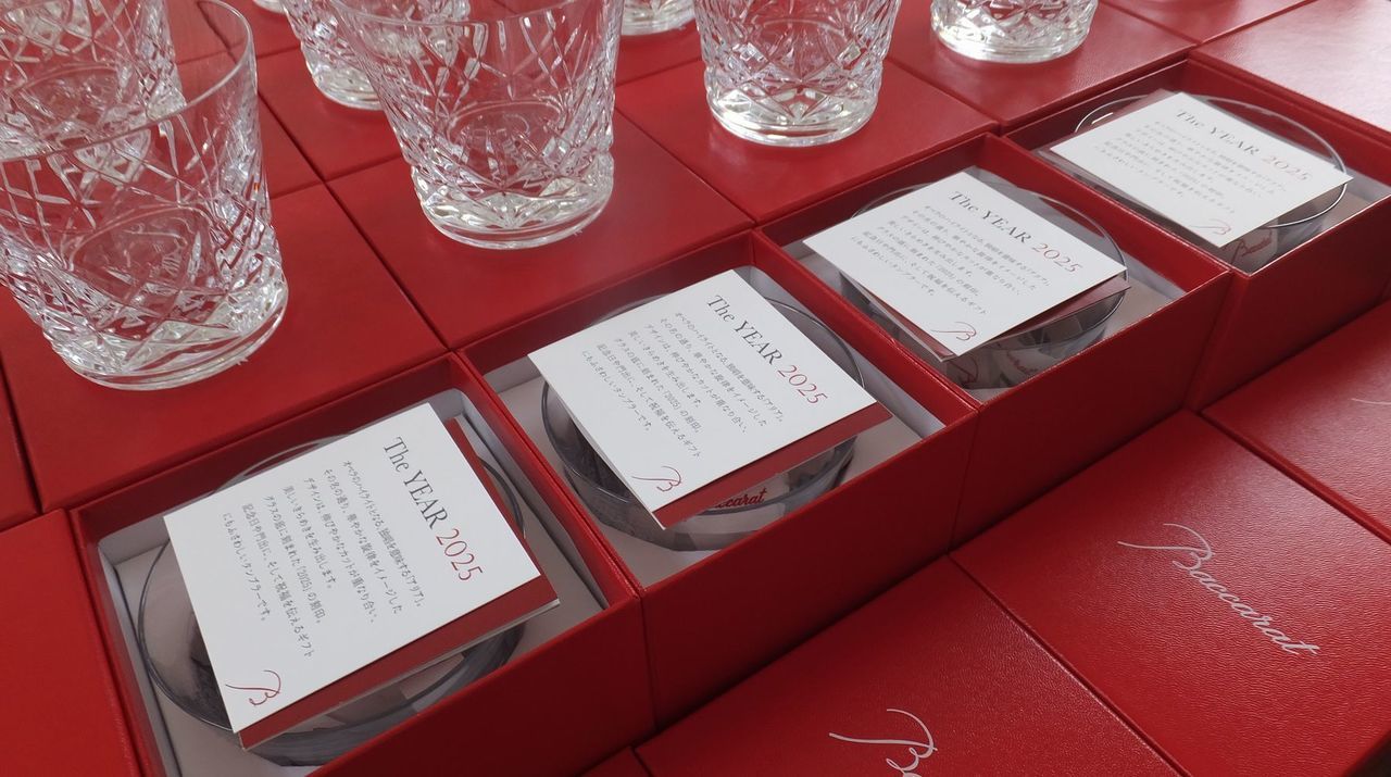 企業の周年祝いを記念して用意されたバカラの2025年最新イヤーグラスが、鮮やかなレッドボックスの上に一つずつ置かれ、画面いっぱいに整然と並べられている圧巻の写真。2025年モデル特有の、未来への希望を感じさせる緻密なカットが光を反射し、無数のクリスタルが宝石のように輝いている。周年を祝う名入れ彫刻が施されたグラスが大量に集まることで、これまでの歩みに対する感謝と、これからのさらなる発展への強い意志を象徴する、壮大で格調高いアニバーサリーシーンを視覚的に表現している。