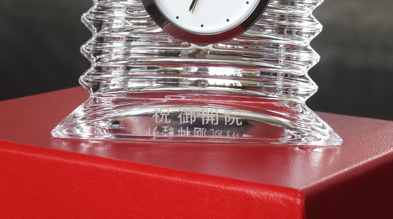 バカラのクリスタル製置き時計『ラランドクロック』に、開院祝いの名入れ彫刻を施した写真。バカラの象徴である鮮やかなレッドボックス（赤箱）の上に置かれた状態で、円形クリスタルの前面下部に『祝 御開院』の文字が、清潔感と信頼を感じさせる格調高い書体で精巧に刻印されている。星のような輝きを放つクリスタルと、ブランドの誇りを象徴する赤いボックスのコントラストが、新しい医療の門出を祝う最高水準の品格と、地域に根ざし時を刻み続ける祝福の気持ちを視覚的に表現している。