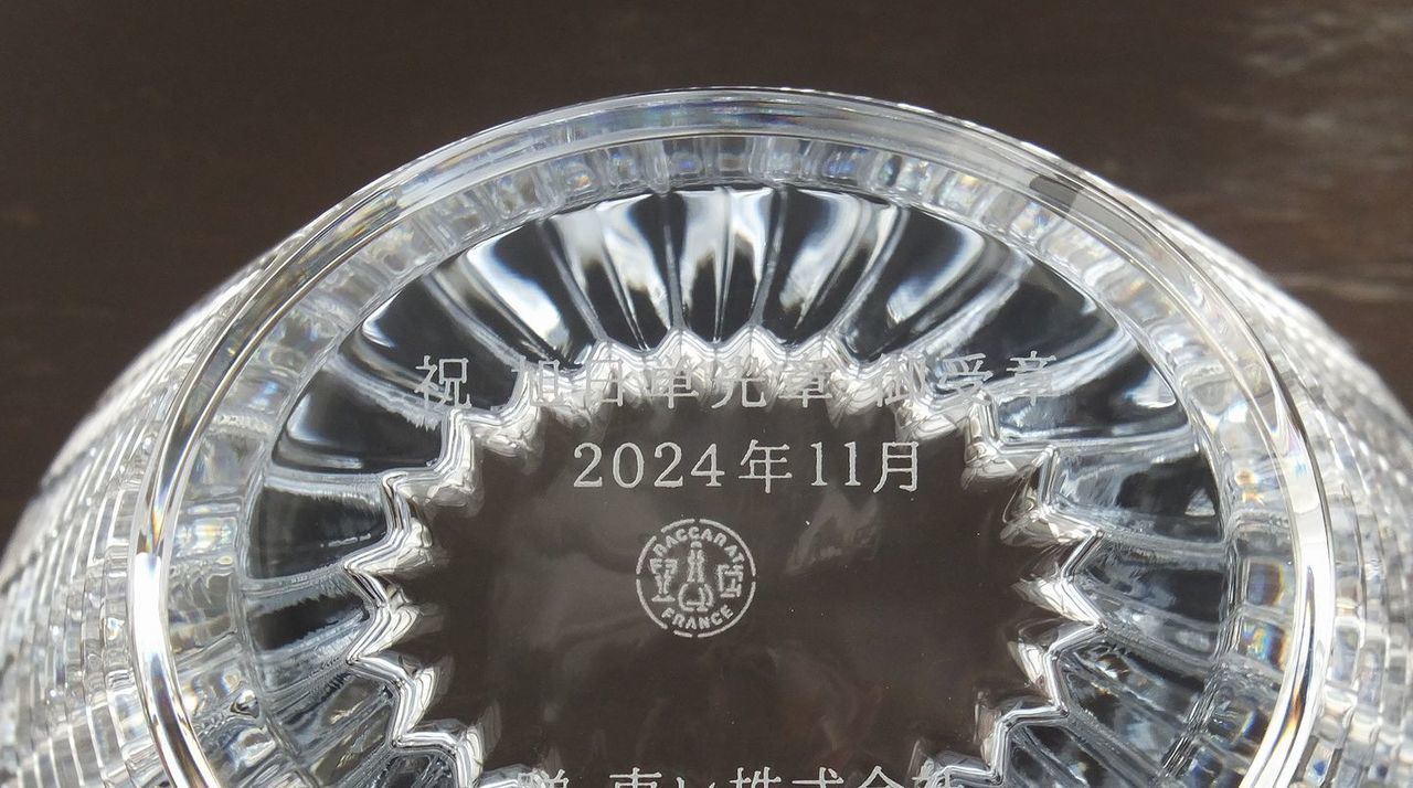バカラのクリスタル製花瓶の側面に、旭日単光章の受章を祝した名入れ彫刻を施したサンプル画像。滑らかなクリスタルの面に、『祝 旭日単光章 御受章』『2024年11月』、そして**『贈り主名』が、格式高く、誠実さを感じさせる楷書体で精緻に刻印されている。バカラ特有の光を美しく透過・反射する重厚なクリスタルが、国家的な功労を称える最高の栄誉を象徴しており、受章者の長年の献身に対する深い敬意と、贈り主からの真心を込めた、気品あふれる一生ものの記念品であることを視覚的に表現している。