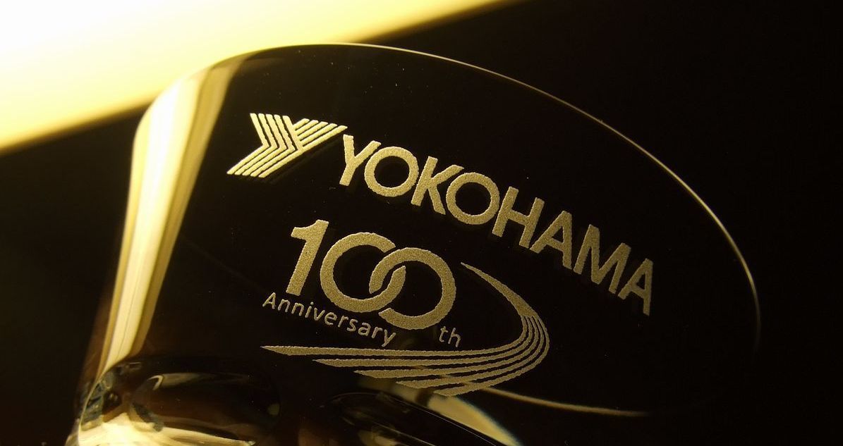 バカラのイヤーグラス（ルチア）のクリスタルグラスのクローズアップ写真。グラスの側面中央に、『ヨコハマタイヤ100周年記念』を祝う特別なデザインが名入れ彫刻されているのが鮮明に確認できる。デザインには、ヨコハマタイヤのロゴ、『100th Anniversary』の文字、そして象徴的な数字『100』が組み込まれ、企業の歴史と発展を称えている。透明度の高いクリスタルに施された精密な彫刻が、企業の品格と未来への展望を象徴する格式高い法人ギフト。