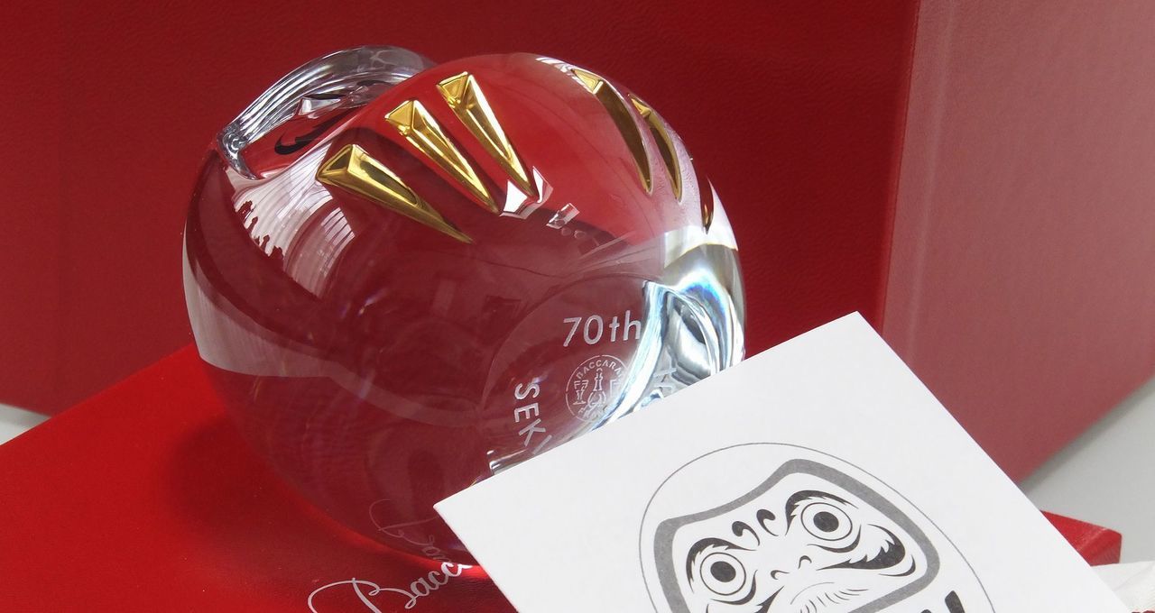 バカラ（Baccarat）のクリスタル製『だるま』の底面に、『70th』の周年記念文字と企業名が精緻に名入れ彫刻された写真。最高級クリスタルならではの輝きと縁起の良いダルマの佇まいに、70周年の節目を祝う刻印が美しく施され、事業のさらなる飛躍と千客万来・商売繁盛を願う周年記念品・法人ギフトの最高峰にふさわしい格調高いイメージ。