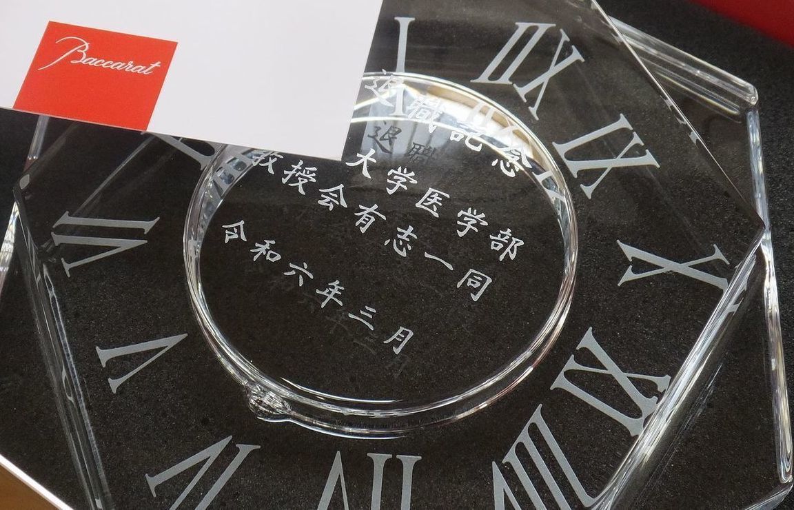 バカラの『アビス クロック』の背面に名入れ彫刻を施した退職記念品の制作実績。「深海」を意味する名前の通り、重厚なクリスタルの塊とエッジの効いたフラットカットが特徴的な置き時計の背面に、『退職記念 [大学名] 医学部 教授有志一同 令和六年三月』の文字が格調高い和文書体で精巧に刻印されている。建築的な美しさを持つクロックに、長年の医学研究と教育への功績を称える送り主一同の想いを刻むことで、圧倒的な存在感と深い敬意を伝える最高峰の記念ギフトとして視覚的に表現されている。