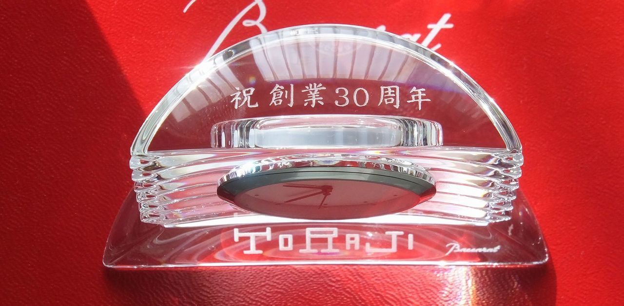バカラ（Baccarat）の置き時計『ラランド クロック』の上部に『祝 創業30周年』、前面下部に『TORAJI』の文字が精緻に名入れ彫刻された創立記念の写真。重厚なクリスタルガラスの質感と眩い円形カットが時を刻む最高級置時計に、企業の30年にわたる歩みと更なる発展への願いを込めた格調高い刻印が光を受けて輝き、会社の周年記念や創業祝い、法人向けのラグジュアリーな記念品に最適なメモリアルギフトイメージ。