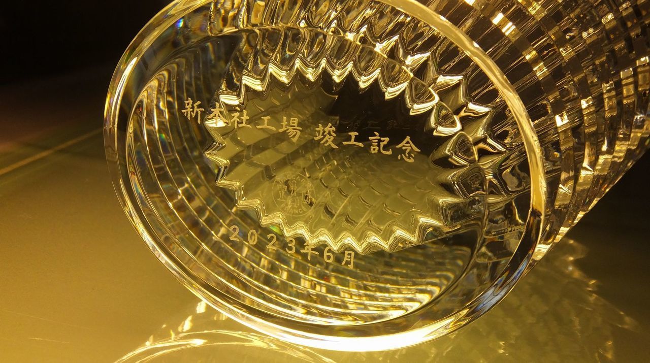 バカラ（Baccarat）のクリスタル花瓶『アイ ベース』の底面に、漢字で『新本社工場 竣工記念』と精緻に名入れ彫刻された写真。独特のカットが光を反射し圧倒的な輝きを放つクリスタルベースに、新たな拠点・新社屋の完成と更なる事業拡大を祝う刻印が美しく施され、企業の新たな飛躍と永続的な繁栄をたたえる最高峰の竣工祝い・法人お祝いギフトにふさわしい格調高いイメージ。