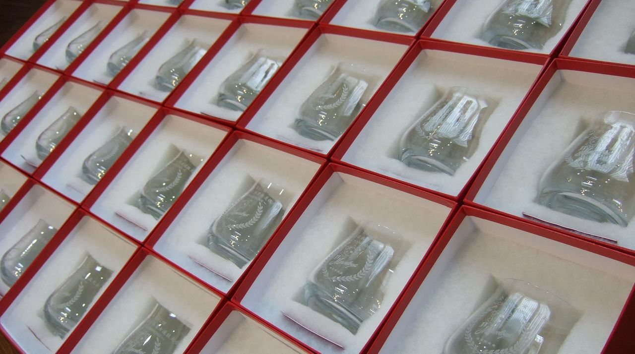 第67回全日本選抜バレーボール大会の優勝を記念して制作された、シャトーバカラ タンブラーが30個以上並ぶ圧巻の光景。各グラスの側面には、『JTマーベラスのチームロゴ』と『第67回大会名』が精緻に彫刻されており、それらがバカラを象徴する鮮やかなレッドボックスの中に整然と収められている。光を反射して輝く大量のクリスタルと、情熱的な赤いボックスのコントラストが、チーム一丸となって勝ち取った栄光の重みと、組織全体の祝祭感を象徴しており、選手や関係者へ贈られる最高峰のチャンピオンシップ・ギフトであることを視覚的に表現している。