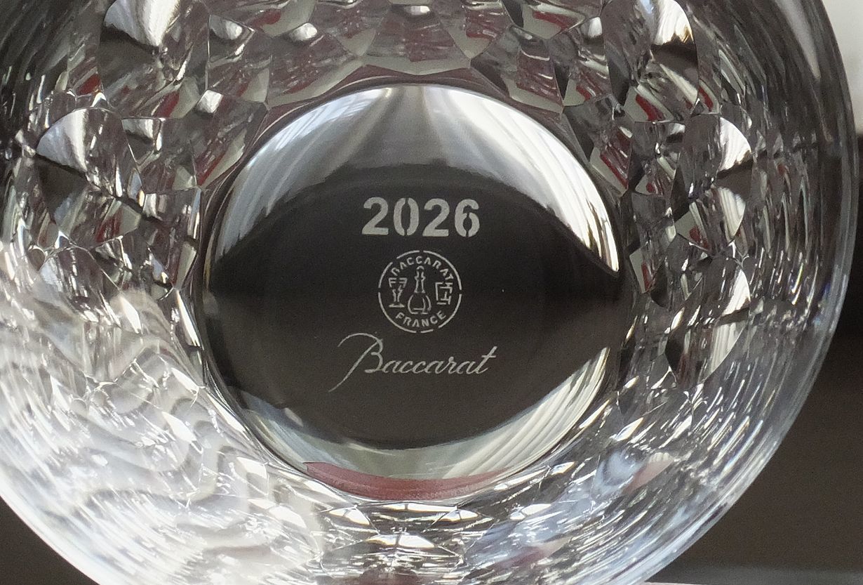 バカラのイヤーグラス、ダリア（Dahlia）タンブラー2026のクリスタルグラスが、テーブルの上に横に倒して置かれ、その底面（フット）を捉えたクローズアップ写真。底面には、『2026』という西暦と、『Baccarat』のロゴマークが繊細に刻印（エッチング）されているのが鮮明に見える。ダリアの花弁を思わせる複雑なカットが施されたグラスの底面が強調され、限定のイヤーグラスとしての確かな証とブランドの格調高さを伝える情景。