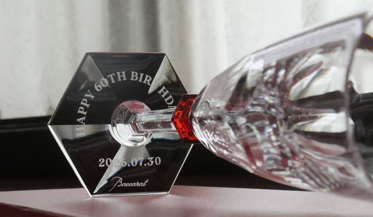 バカラ（Baccarat）の『アルクール イヴ シャンパングラス』の底面に、『Happy 60th Birthday』のメッセージと記念日付が精緻に名入れ彫刻された、ファンクラブからアーティストへ贈られた還暦祝いの写真。バカラのアイコンである『アルクール』をよりしなやかでエレガントに進化させた最高級クリスタルグラスを通し、繊細なフット（底面）から溢れる感謝と情熱を込めた言葉が密やかに光を浴びて輝き、特別な節目や愛するアーティストの長寿・メモリアルを祝う最高峰のオーダーメイドギフトイメージ。