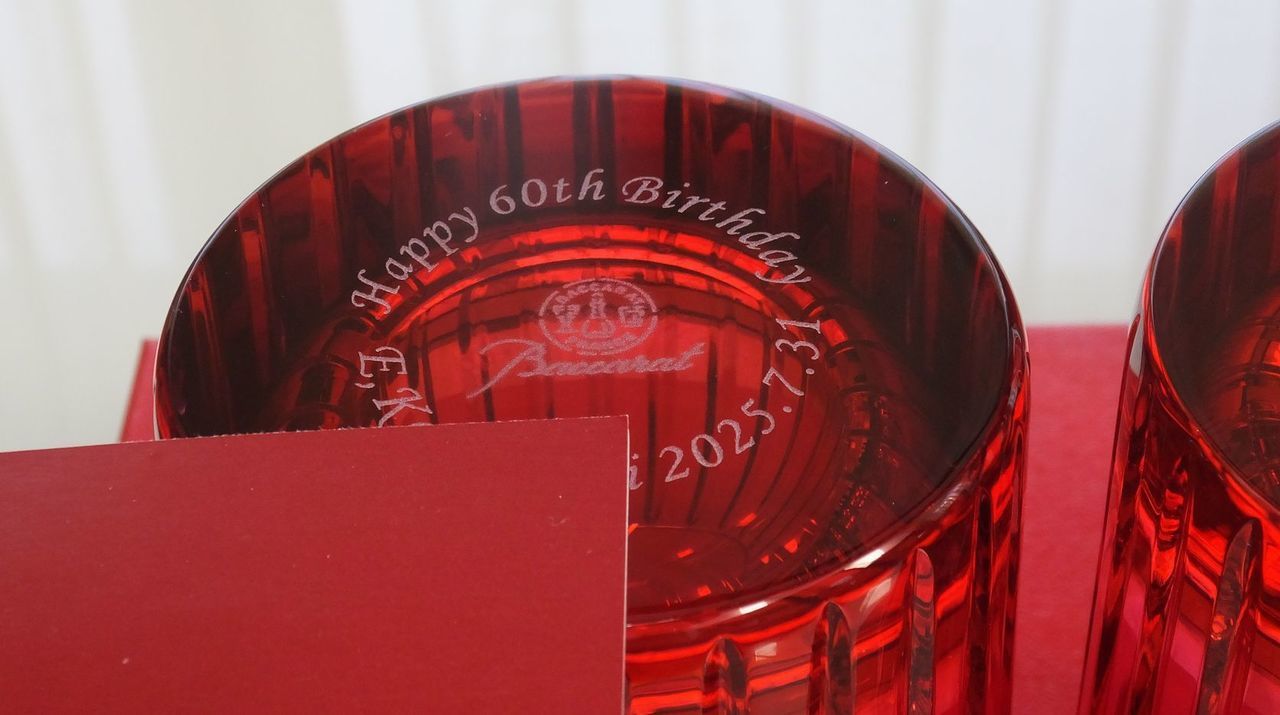 バカラ（Baccarat）の赤いクリスタルタンブラー『ハーモニー レッド』の底面に、英語で『Happy 60th Birthday』のメッセージ、お名前、および日付が精緻に名入れ彫刻された写真。鮮やかなレッドのクリスタルと垂直のカットラインが美しい光を放つグラスに、還暦という特別な人生の節目を祝福する刻印が映え、60歳の誕生日祝いや還暦祝い、最高峰のパーソナルギフトにふさわしい格調高いイメージ。
