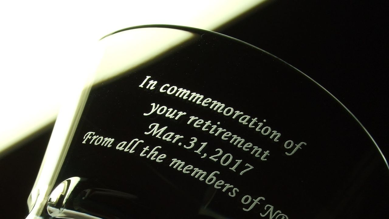 バカラのイヤータンブラー（年号刻印入り）の側面に、格調高い英文フォントで『In commemoration of your retirement』とお名前、日付を名入れ彫刻したサンプル写真。底面に刻まれた年号と、側面の退職記念メッセージが重なり合い、長年の献身的な歩みへの深い敬意と感謝を優雅に表現する、最高級の法人向け定年退職・門出祝いギフトのイメージ。