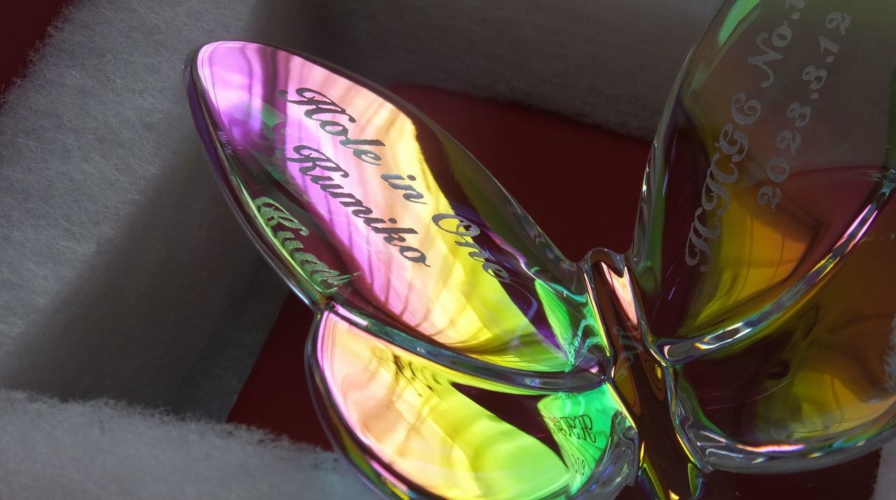 バカラのクリスタルオブジェ『ラッキーバタフライ イリゼクリア』の両方の羽根に、ホールインワン達成の記念彫刻を施したサンプル画像。左の羽根には『Hole in One』と『達成者のお名前』、右の羽根には『ゴルフ場名・コース名』と『達成日付』が、軽やかで優雅な筆記体フォントで精緻に刻印されている。光の加減で虹色に輝くイリゼ（虹色）加工が、まさに奇跡とも言えるホールインワンの歓喜と幸運を象徴しており、ゴルフ人生における最高の瞬間を永遠に輝かせる、繊細で美しい特別な記念品であることを視覚的に表現している。