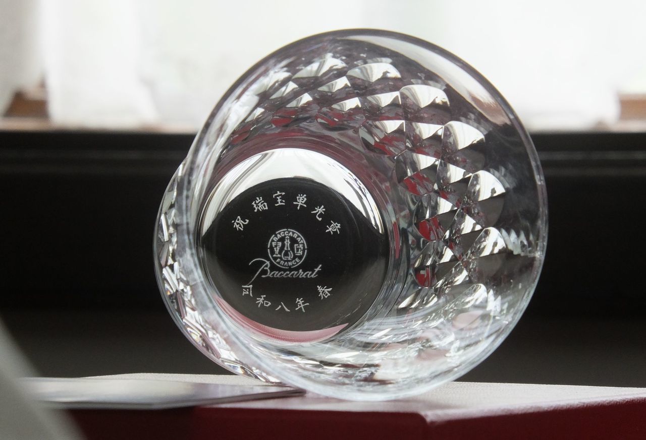バカラ（Baccarat）の『アルマニャック タンブラー』の底面に、『祝 瑞宝単光章 令和八年 春』の文字が精緻に名入れ彫刻された写真。ダイヤ状のカットが交差し華やかな光を放つ最高級クリスタルグラスの底から、国家的な受章の栄誉と長年の貢献をたたえる刻印が密やかに美しく輝き、叙勲・褒章の受章記念品や特別な格調高い法人ギフトにふさわしい最高峰のイメージ。