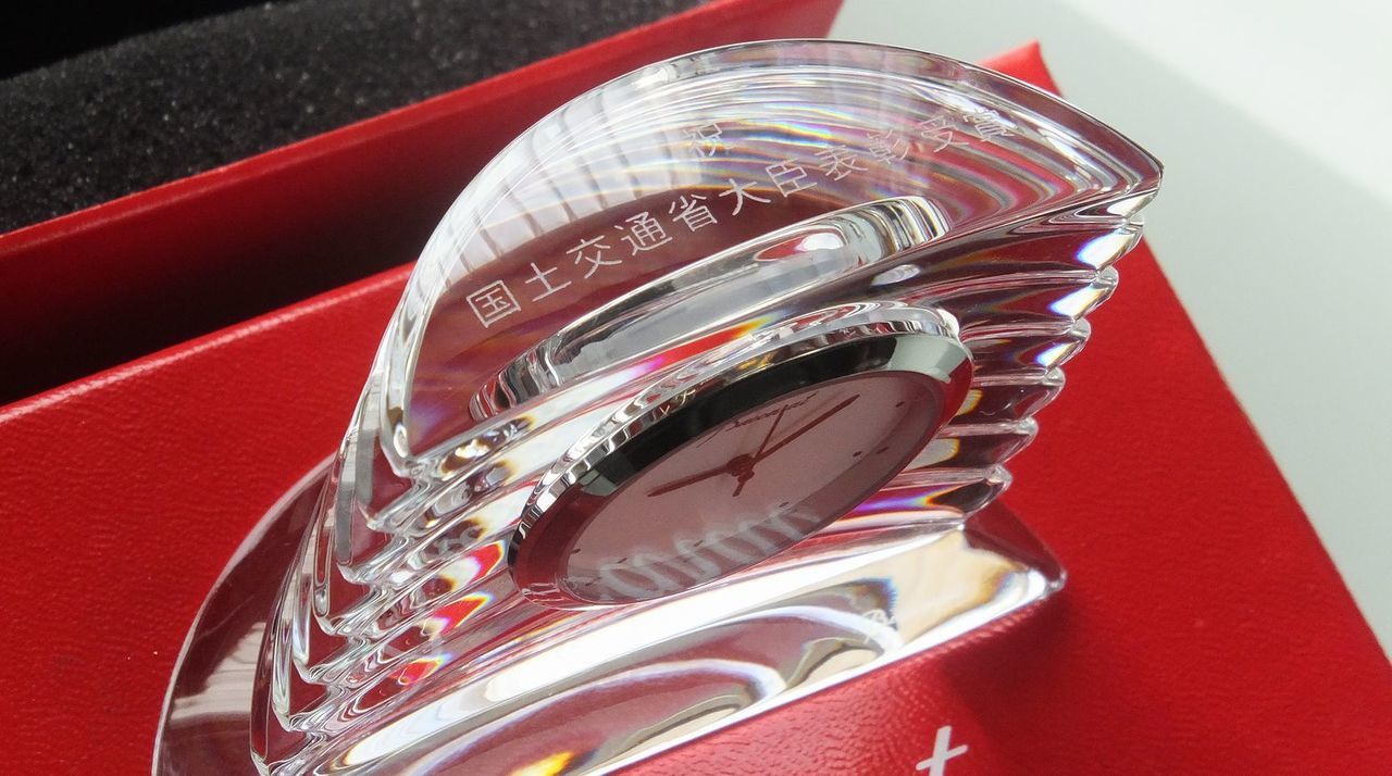 バカラのフラワーベース『ギンコ（銀杏）』に、東証プライム上場記念の名入れ彫刻を施した逸品。扇形に広がる銀杏の葉をモチーフにしたエレガントなフォルムの側面に、精緻な企業ロゴとともに『祝 東証プライム上場 [企業名] 2025』の文字が、重厚で格式高い書体により精巧に刻印されている。「不老長寿」や「繁栄」の象徴である銀杏をデザインしたこの花瓶は、企業の新たな歴史の幕開けと永続的な発展を祝うにふさわしい、圧倒的な気品と輝きを放つ最高峰の記念ギフトであることを視覚的に表現している。