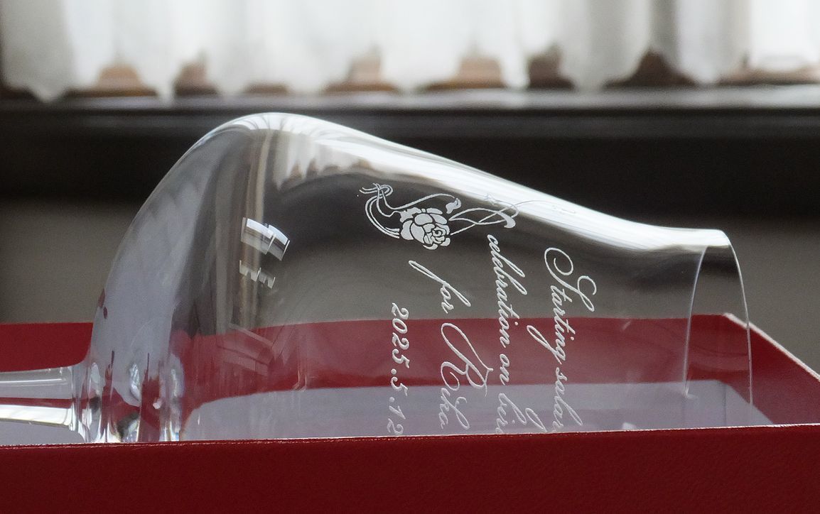 バカラ（Baccarat）の『シャトーバカラ ワイングラス』の側面に、優美なバラ（薔薇）のデザインモチーフとともに2行のお祝いメッセージ、お名前、日付が精緻に名入れ彫刻された開業祝いの記念写真。ワインの芳醇な香りを引き立てる洗練されたモダンなフォルムと最高級クリスタルグラスの透明感に、華やかなバラのカットと門出を祝う刻印が美しく映え、クリニックやサロンの開業祝い、店舗のオープン記念、企業創立の特別なギフトにふさわしい格調高いイメージ。