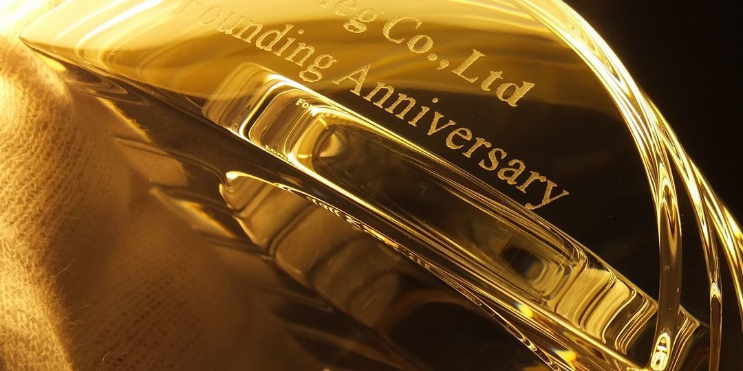 バカラ（Baccarat）のクリスタル製置時計『ラランドクロック』の上部に、『設立記念』『〇〇株式会社』といった設立記念の英語の文字と企業名が精緻に名入れ彫刻された写真。最高級クリスタルならではの圧倒的な透明感と輝きを放つ時計に、企業の永続的な発展と信頼を刻んだ特別なギフト・記念品。デスクやエントランスを格調高く彩る、洗練されたイメージ。