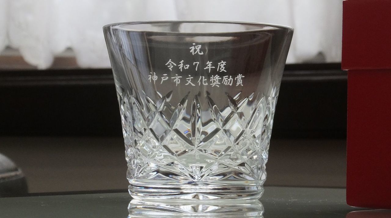 バカラのクリスタルグラスの側面に、令和7年度の文化奨励賞受章を祝した名入れ彫刻を施したサンプル画像。グラスの表面に**『祝 令和７年度 神戸文化奨励賞』の文字が、芸術的感性と気品を感じさせる美しい書体で精巧に刻印されている。バカラ特有の澄み切った輝きが、地域文化の発展に寄与した功績と、今後のさらなる創造的な活躍を象徴しており、受章者のたゆまぬ努力と才能を称える、一生の宝物にふさわしい格調高い記念品であることを視覚的に表現している。
