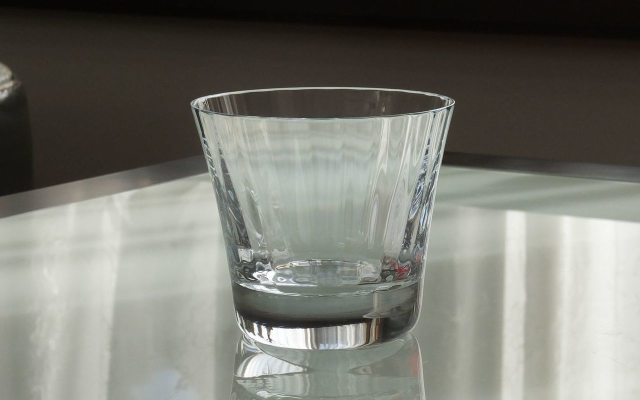 バカラの『ミルニュイ タンブラー』が、モダンなガラステーブルの上に置かれている様子の写真。アラビアンナイトをイメージしたミルニュイ特有の、プリーツ状のカットが光を受けて美しくきらめいている。透明なガラステーブルの上では、タンブラーのクリスタルカットの輝きが反射し、光と影のコントラストを際立たせ、ラグジュアリーで洗練された雰囲気を醸し出している。