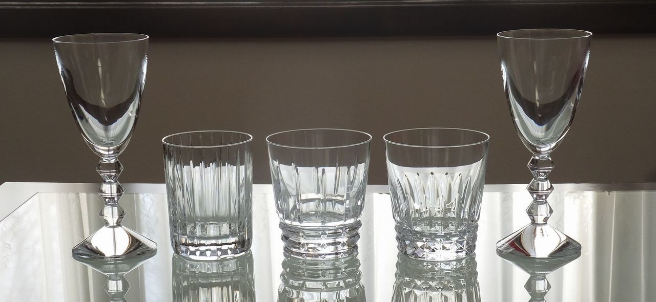 4つのバカラ製タンブラー（アビス、アルマニャック、ローハン、アルルカン）が、モダンなガラステーブルの上に横一列に並んでいる写真。左から、重厚なフラットカットが特徴のアビス、斜めの深いカットが輝くアルマニャック、繊細な蔓草模様のエッチングが施されたローハン、そして色彩豊かな道化師の衣装をイメージしたカットのアルルカンが並び、それぞれのクリスタル特有の輝きとデザインの個性が際立っている。透明なガラステーブルが光を透過させ、各グラスの複雑なパターンと美しい影を美しく映し出し、バカラの多様な芸術性を一堂に感じさせる洗練された光景を表現している。