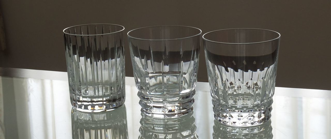 バカラのクリスタルグラス3種、ハーモニータンブラー、アルルカンタンブラー、ティアラタンブラーが、テーブルの上に並べて置かれている集合写真。縦のラインが特徴的な『ハーモニー』、ひし形（アルルカン）のカットが特徴的な『アルルカン』、そして繊細なティアラ状のカットが特徴的な『ティアラ』が、それぞれ異なる光の反射と輝きを見せている。バカラクリスタルの多様なカット技術とデザインの美しさを伝える、コレクションやギフトの比較に適した情景。