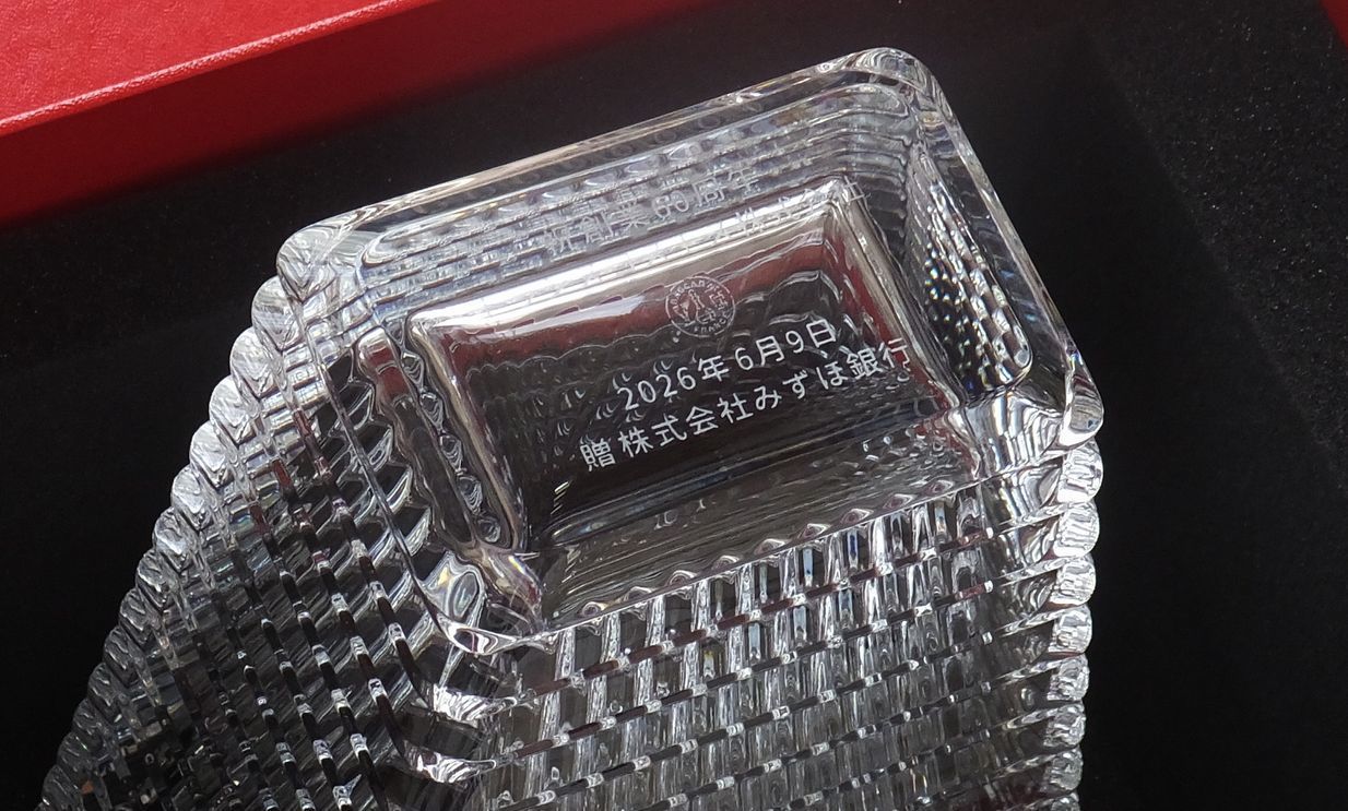 バカラ（Baccarat）のクリスタル花瓶『アイ ベース スクエア』の底面に、漢字で『2026年6月9日 贈 株式会社みずほ銀行』と精緻に名入れ彫刻された写真。独自のウェーブカットが光を美しく反射する最高級クリスタルならではの重厚感と透明感に、企業の記念日や節目を祝う上品な刻印が映え、法人向けの創立・周年記念品、就任祝い、上場祝いなどの大口・高級贈答品にふさわしい格調高いイメージ。