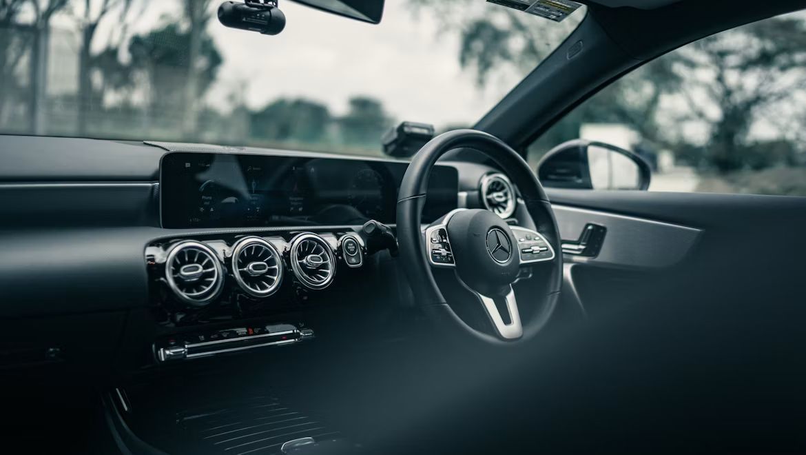 メルセデス・ベンツ（Mercedes-Benz）の車内にて、後部座席から運転席・コックピット方向を見渡した写真。洗練されたステアリングホイール、先進的なデジタルディスプレイやインパネ周り、上品なインテリアライティングが広がり、ドイツ高級車ならではのラグジュアリーな室内空間と優れた居住性、快適なドライブ体験を演出するイメージ。