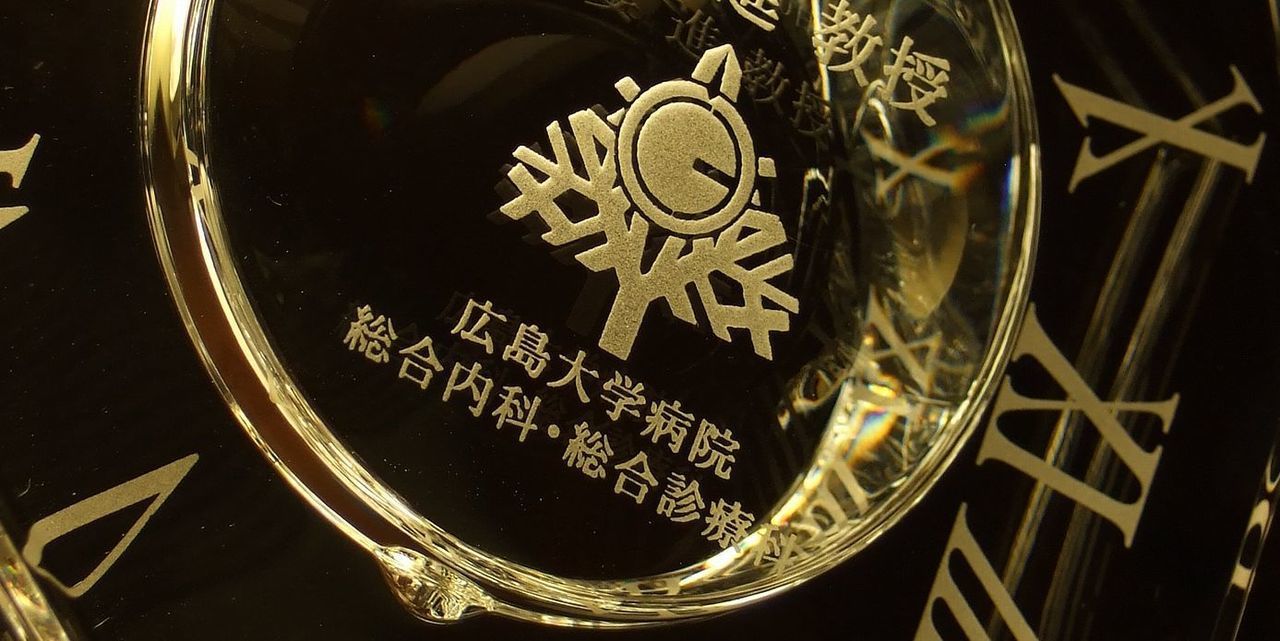 バカラ（Baccarat）の『アビス』クロックの背面に、広島大学病院のロゴマークデザインが精緻に名入れ彫刻された写真。深みのある重厚なバカラクリスタルのフォルム越しに、精密に刻まれたロゴデザインが立体的に浮かび上がり、病院の周年記念、教授就任・退職記念、新病院棟の開院など、高度医療機関における節目と功績を称える特別で格調高い記念品イメージ。