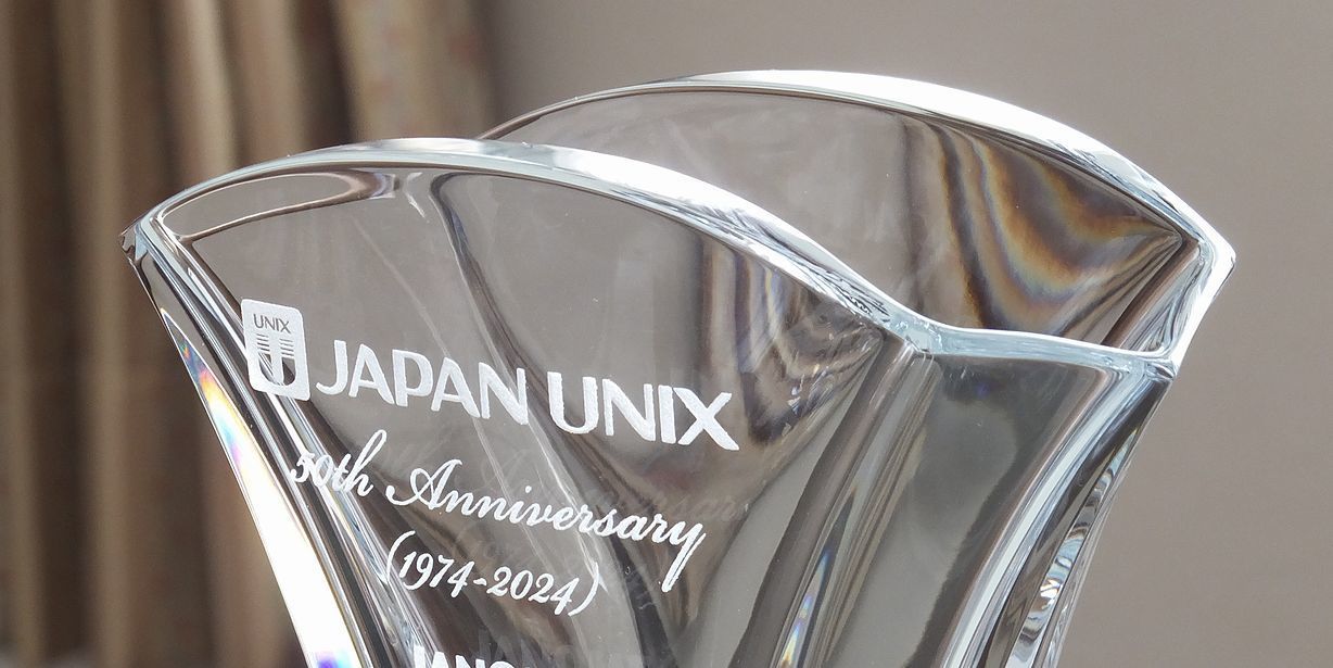 バカラのギンコベース（花瓶）の側面に『JAPAN UNIX 50th Anniversary』というメッセージと企業ロゴを名入れ彫刻を施した写真。銀杏の葉をモチーフにした優美なクリスタル製花瓶に、企業の50周年を祝うメッセージとシンボルマークが丁寧に刻まれています。企業の輝かしい歴史と未来への発展を象徴する、格調高い特別な周年記念ギフトです。