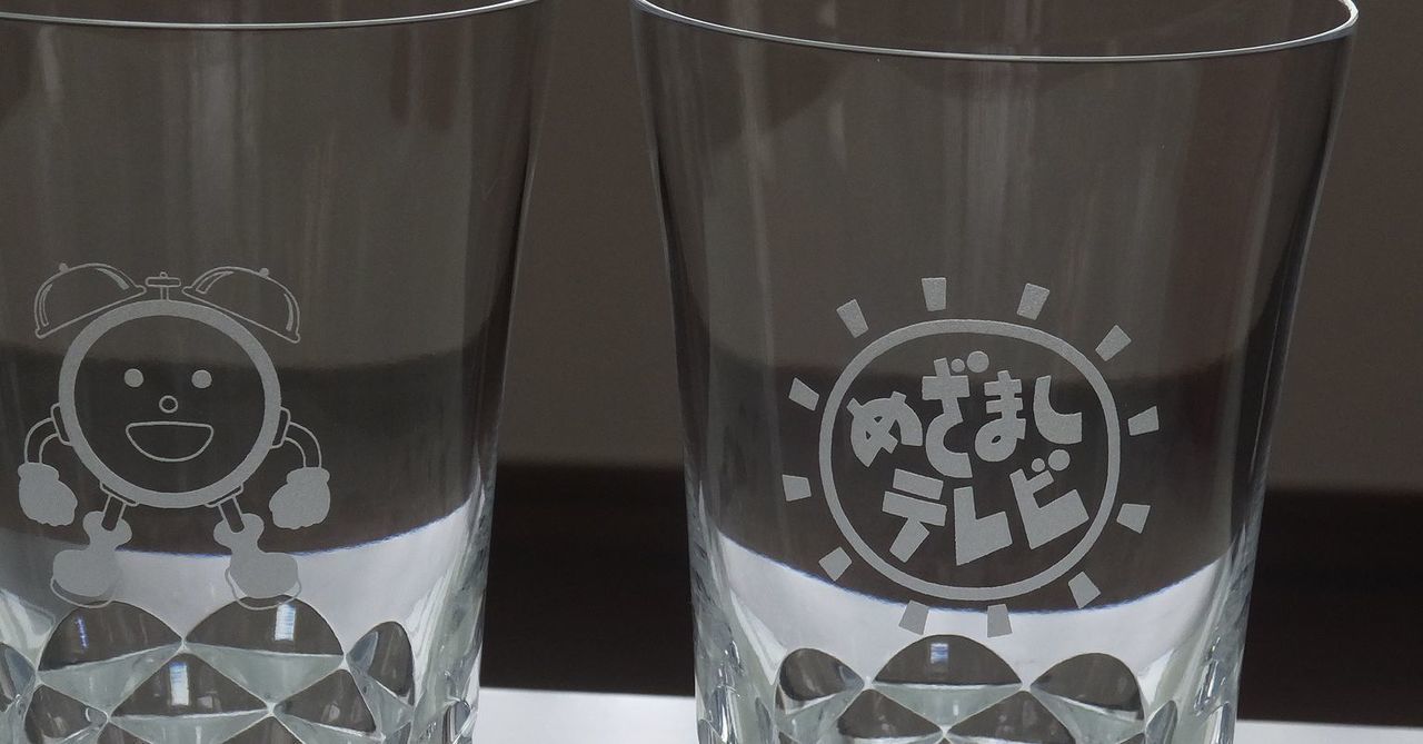 バカラの『ブラーヴァ ハイボール』ペアグラスに、めざましテレビ卒業記念の特注彫刻を施した贈り物。喝采（ブラーヴォ）を意味する名を持つ、扇を広げたようなカットが美しいグラスの側面に、『めざましテレビ』の番組ロゴと、愛らしいキャラクター『めざましくん』が精巧に刻印されている。番組を卒業するキャスターへの長年の感謝と門出を祝う拍手が込められており、クリスタルが放つ輝かしい光が、朝の顔として活躍した日々への敬意を象徴する世界に一つだけの特別な記念品であることを視覚的に表現している。