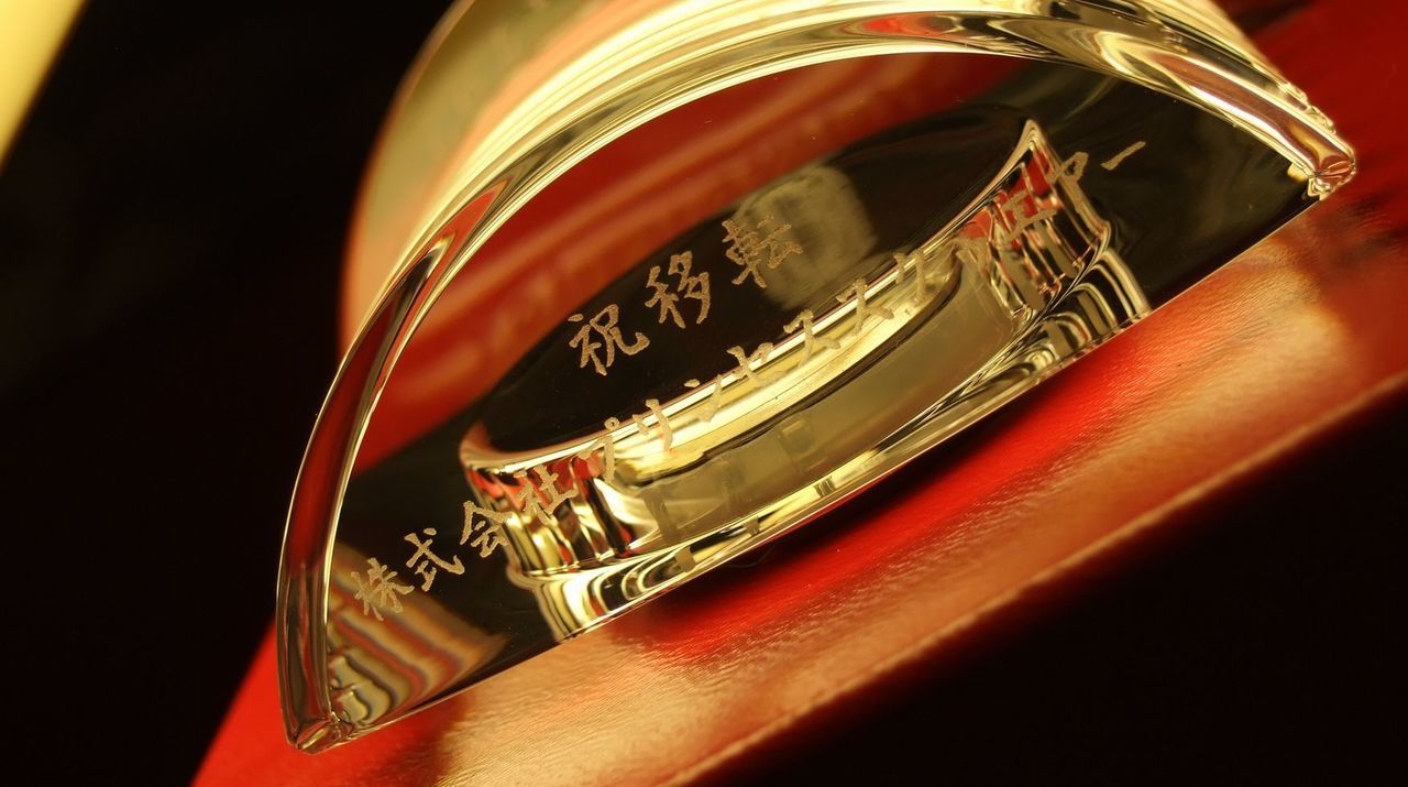 バカラのクリスタル製置き時計『ラランドクロック』の前面上部に施された、移転祝いの彫刻のアップ写真。円形クリスタルの縁に沿って、『祝移転 [社名]』という文字が、凛とした高級感のある書体で深く鮮明に刻印されている。クリスタルの透明な層越しに光が透過し、彫刻された文字が白く美しく浮かび上がっている。新天地での新たな時の刻みを象徴するこの時計が、移転という重要な節目を称える最高級の贈答品であることを、質感まで伝わる高精細な接写で表現している。