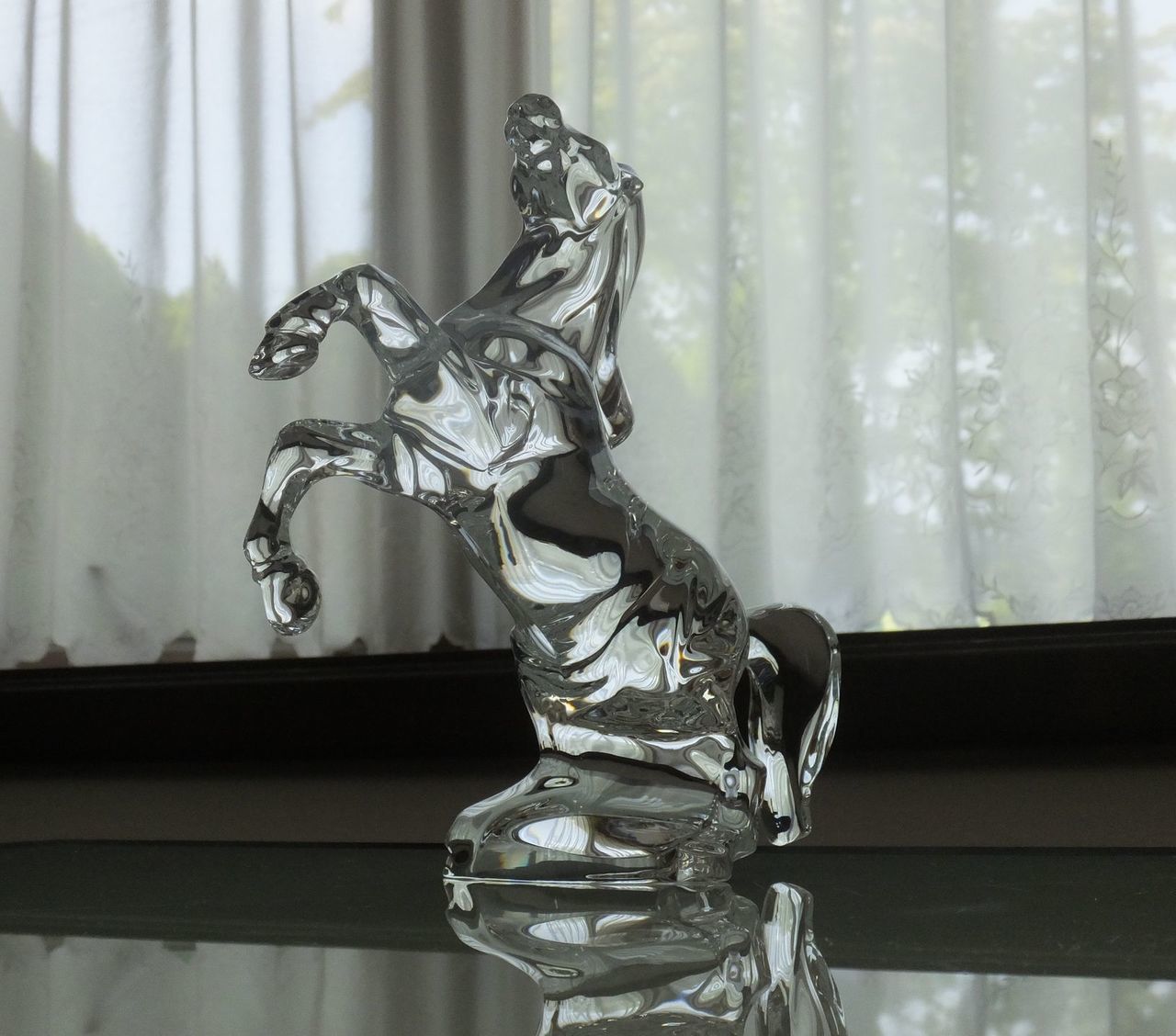 洗練されたテーブルの上に置かれた、バカラ（Baccarat）のクリスタル製オブジェ『いななく馬』の写真。前足を高く上げ、今にも動き出しそうなほど生命力と躍動感にあふれる美しいシルエット。最高級クリスタルガラスならではの圧倒的な透明感と滑らかなカットが光を均一に反射し、空間に極上の輝きと品格をもたらす。栄誉や成功、躍進を象徴する格調高いフィギュリンとして、特別な記念日や法人向けの最高峰のギフトにふさわしいラグジュアリーなイメージ。