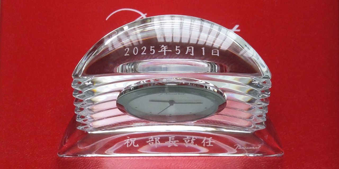 バカラ（Baccarat）のクリスタル製置時計『ラランドクロック』の上面に日付、前面下部に漢字で『祝 部長就任』の文字が精緻に名入れ彫刻された写真。最高級クリスタルならではの透き通る輝きと重厚な存在感を放つ時計に、組織での新たなる役職への昇格と更なるご活躍を祝福する刻印が美しく施され、部長就任祝いや役員・幹部昇進のお祝いにふさわしい最高峰の法人贈答ギフト・記念品のイメージ。